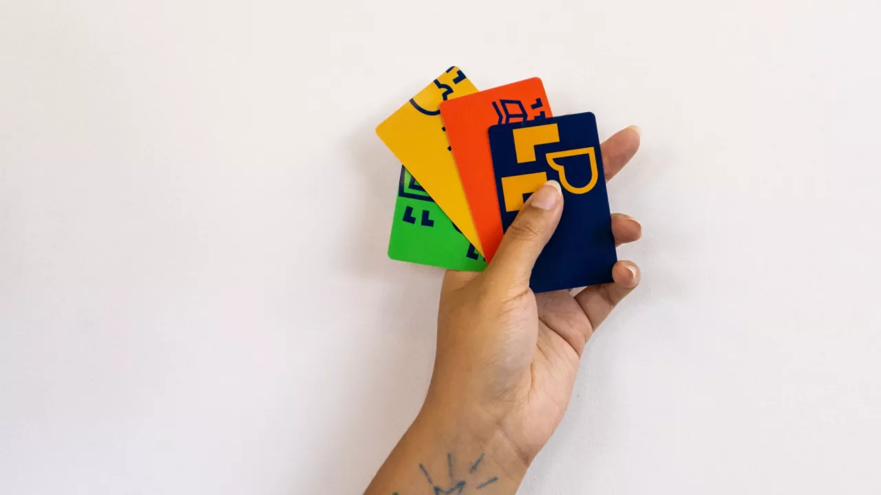 A photo of a hand holding 4 different coloured London Public Library cards against a white wall. You can see a tattoo on the outstretched arm.