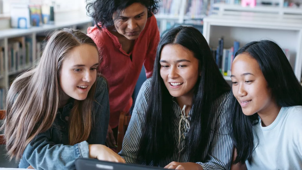 Four older students sit at table looking at laptop and smile.  In the background are bookshelves.