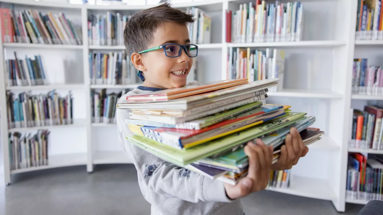 Photo of young boy in glasses holding a large stack of books. In the background are bookshelves.