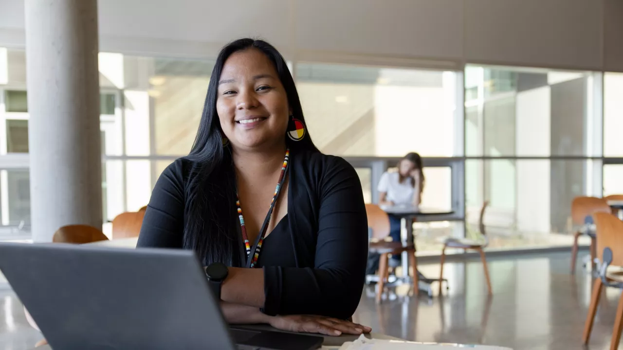 Photo of a young woman sitting at a table in a library. On the table is a laptop. In the background are windows and another person sitting at a table.