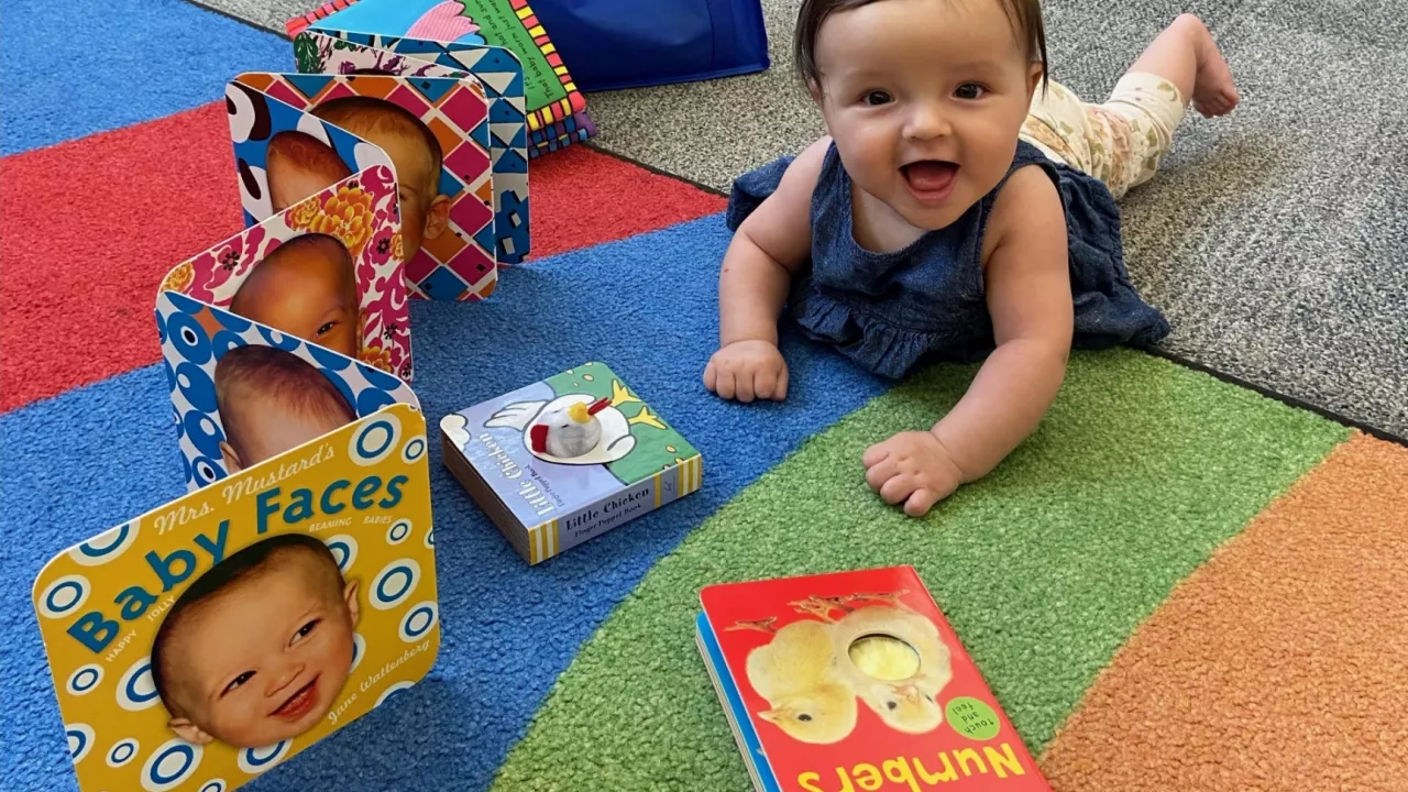 Photo of a baby lying on a carpet in the library looking up and smiling. In front of the baby are several board books.