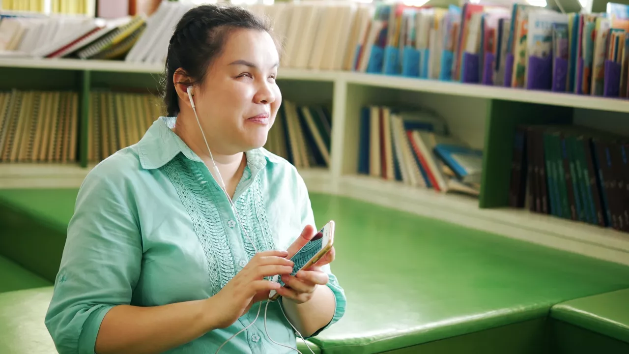 Photo of a woman with low vision using smart phone with earphones connected. In the background are bookshelves. The woman is wearing a green shirt and is leaning against a green desk.