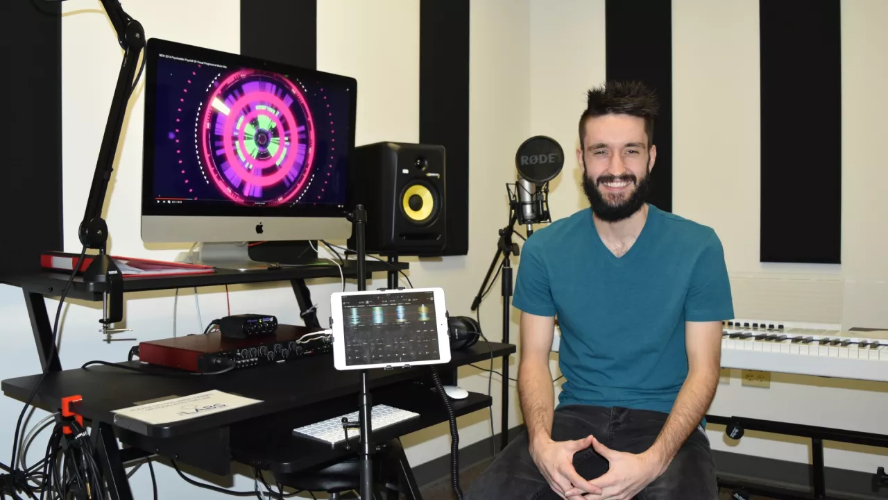 Photo of a smiling man sitting in the audio recording studio. Beside him is a keyboard and monitor. In the back are speakers and in front is a microphone.