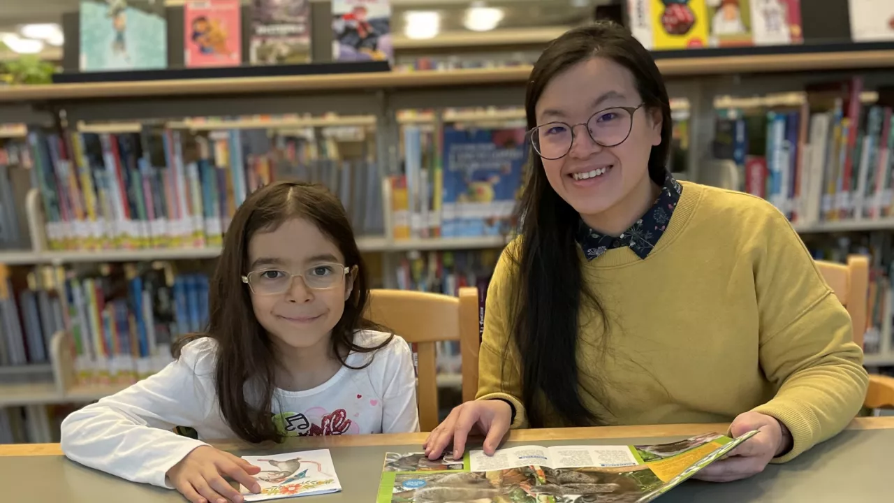 Photo of a child and young woman sitting beside each other at a table and looking up and smiling. They are holding books. In the background are bookshelves.
