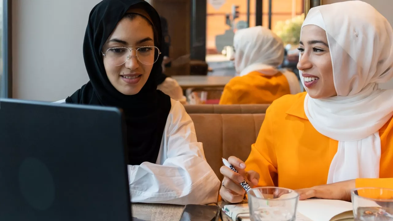 Photo of two women smiling and talking in a cafe. On the table is a laptop and notebooks.
