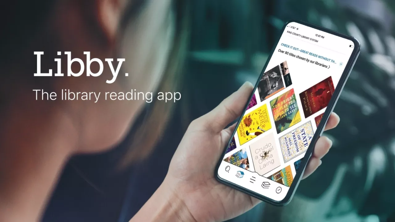 A photo of a youth holding a phone in their hand on which is displayed the Libby app with book covers. Over the photo is the text Libby. The library reading app.