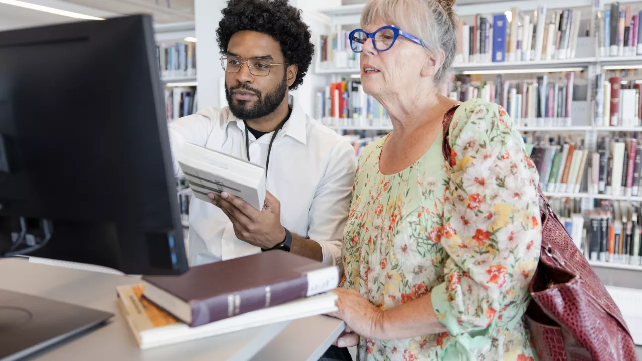 Photo of a male library worker standing with an older, female patron at a computer terminal. They are holding books and looking the screen.