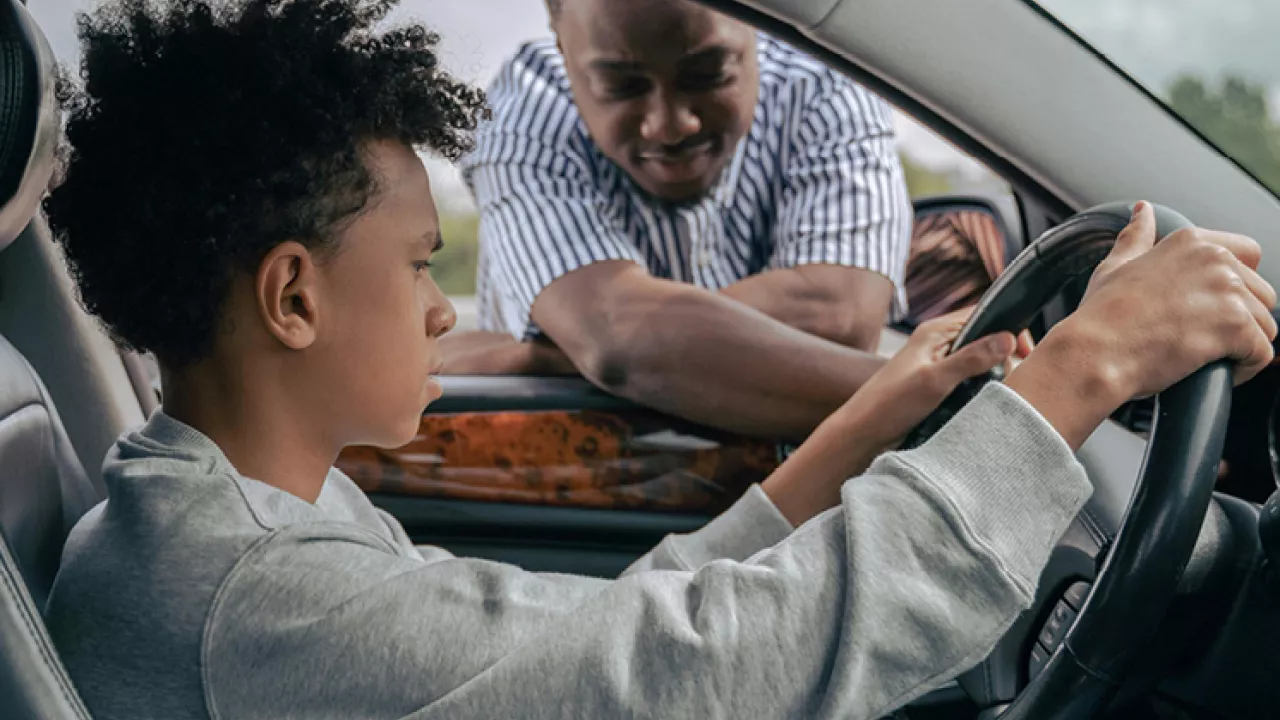 Young person learning to drive with older person leaning in through the driver's window