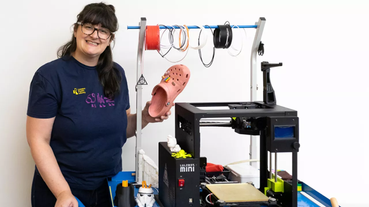 A librarian standing beside a 3D printer smiling and holding up a croc with 3D printed accessories
