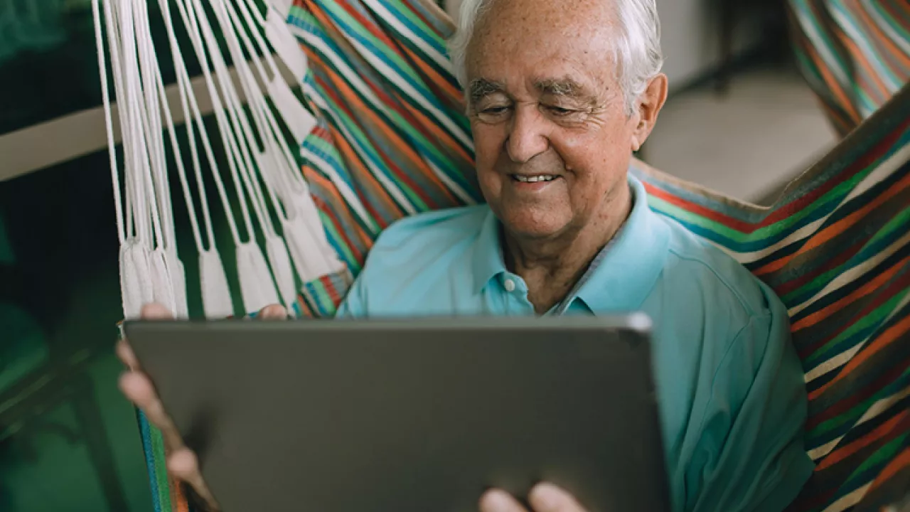 An older man sits in a hammock while reading on a tablet