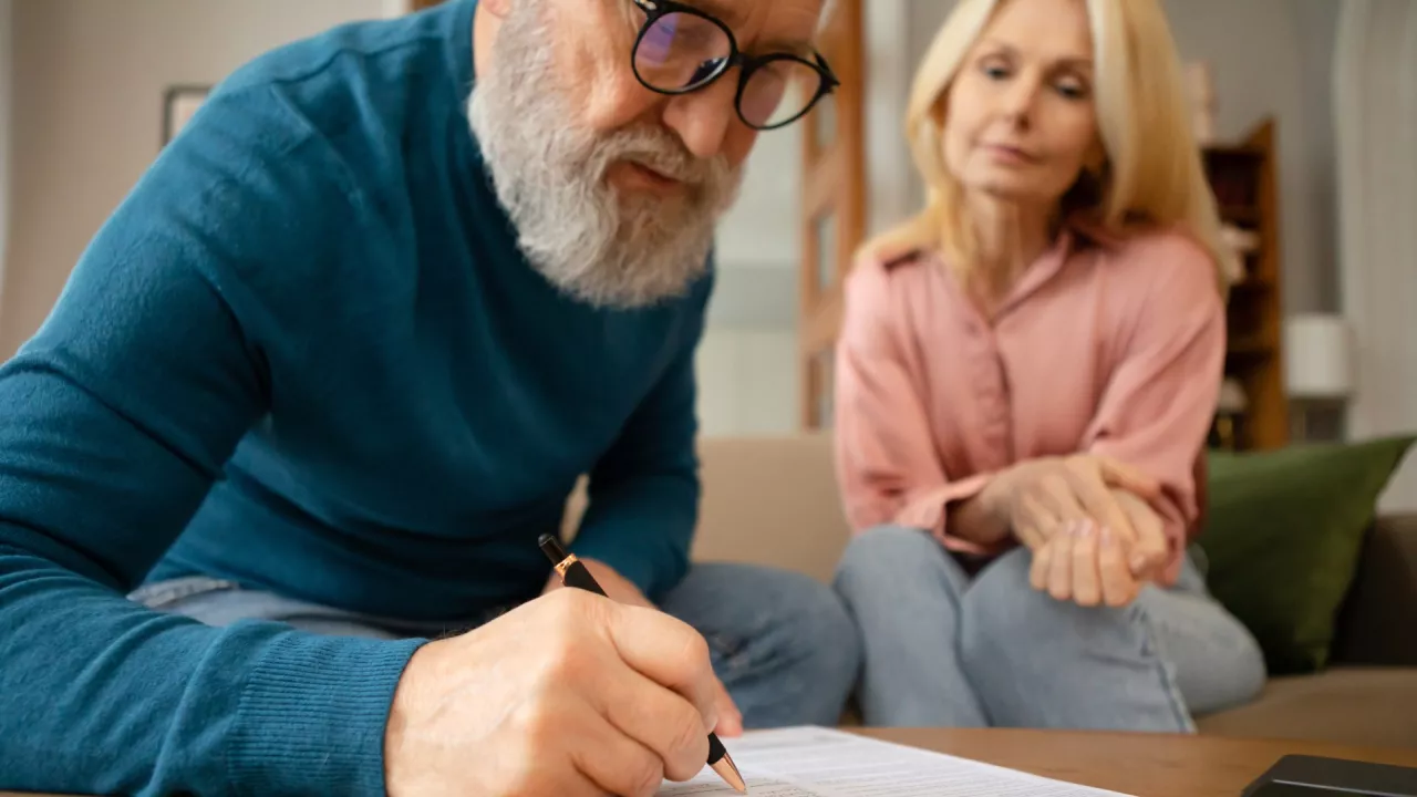 A man in a blue sweater fills out a form with the help of a woman watching