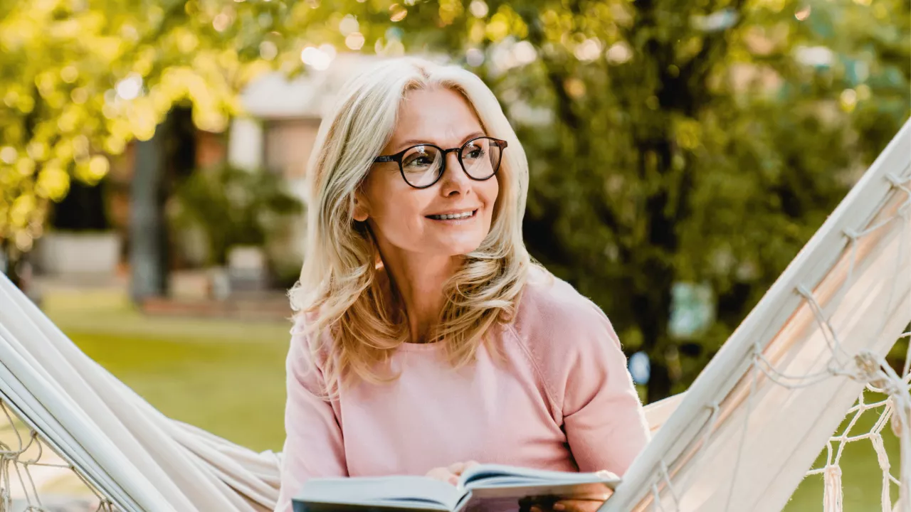 Smiling senior blond woman wearing glasses while reading in hammock in the summer garden