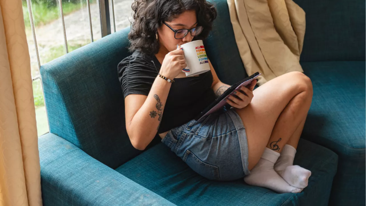 A woman sitting on a blue chair reading on a tablet while drinking coffee