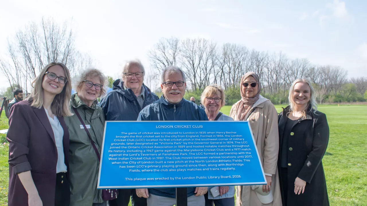 7 people, members of the Historic Sites Committee stand behind a blue plaque. In the background is parkland.