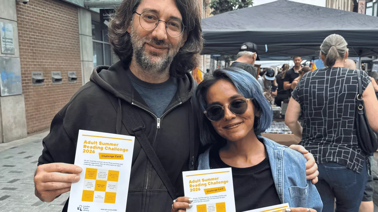 Two adults smile while holding up challenge cards for the Adult Summer Reading challenge.