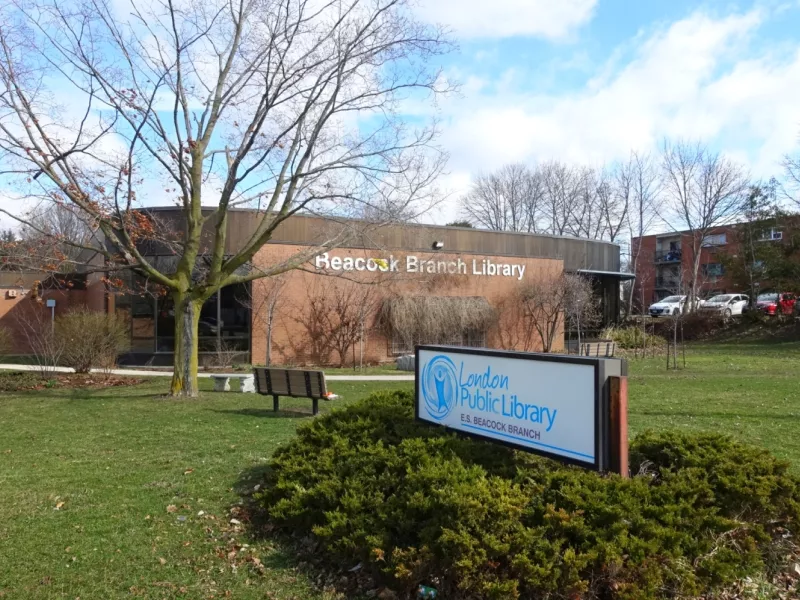 A photo of the outside of Beacock Branch showing the Library sign in front and the building in the back with a tree, bench and shrub on the grass in between.