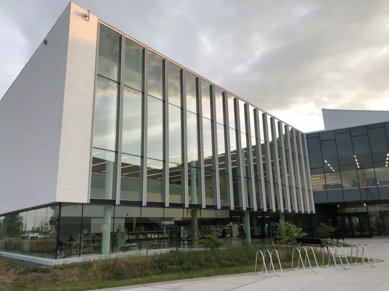 Photo of the outside of Bostwick Branch Library with light reflecting off the glass. Bookshelves can be seen inside and a bike rack sits in front of the building.