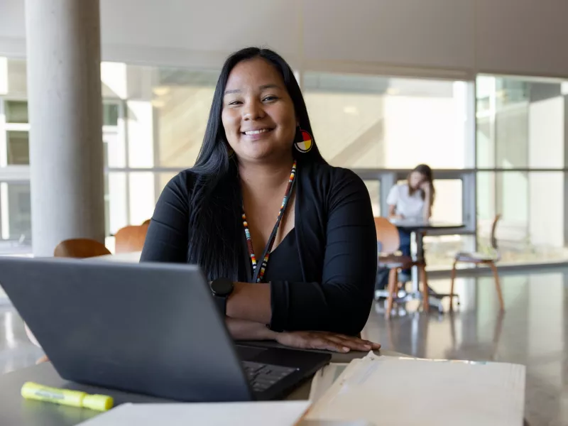 Photo of a young woman sitting at a table in a library. On the table is a laptop. In the background are windows and another person sitting at a table.
