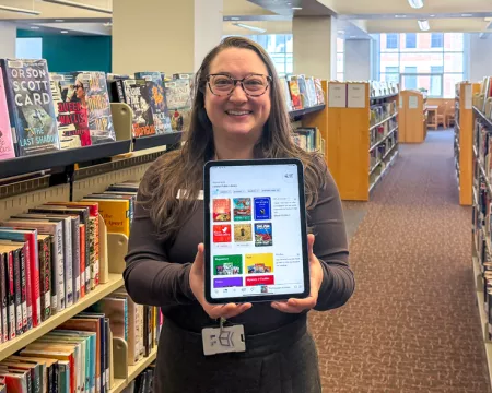 Librarian standing in between book shelves holding out an iPad with the Libby app loaded.