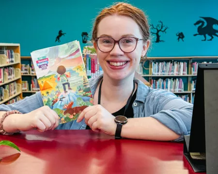 Claire holds up a TD summer flyer in the Children's area, surrounded by bright colourful books.