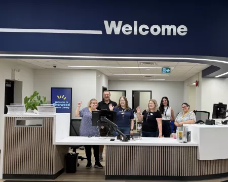 The staff of Sherwood Branch Library smile and wave from behind their new service desk above which is the word Welcome