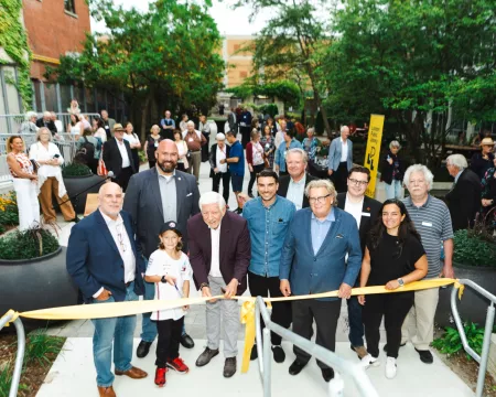 Local dignitaries donors and Library staff gather behind a yellow ribbon as a young boy holding scissors prepares to cut the ribbon in the Rotary Reading Garden.
