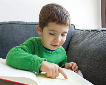A boy about 6 or 7 years old reads a book using his finger to follow the text. 