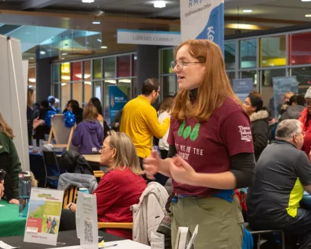 Inside a busy library atrium, community members gather around information tables at an event. In the foreground, a person wearing a maroon T-shirt speaks with an older man holding a camera across a table with pamphlets and a QR code sign. In the background, many people are seated and standing, talking with representatives at different booths, creating a lively, crowded atmosphere.