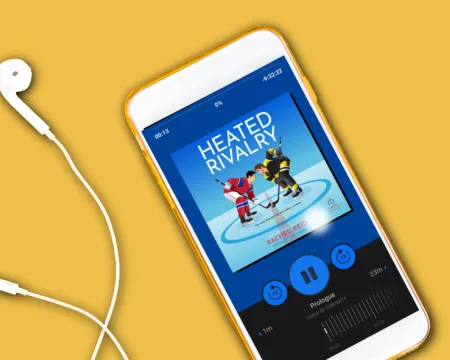 A smart phone with headphone plugged in on a yellow background. The screen shows the audiobook of Heated Rivalry playing.