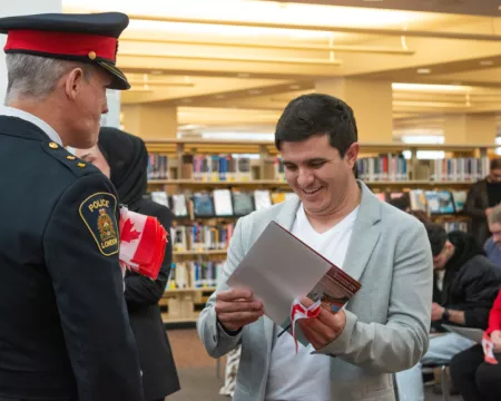 A man holds his citizenship certificate and smiles while a London Police Services Officer in uniform looks on. In the background participants sit on chairs surrounded by bookshelves.
