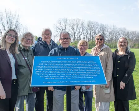 7 people, members of the Historic Sites Committee stand behind a blue plaque. In the background is parkland.