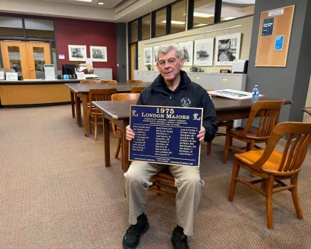 Barry Boughner sits in the London Room at Central Library holding a blue commemorative plaque recognizing the 1975 London Majors championship team. Research tables, archival storage cabinets, and historic photographs line the room behind him.
