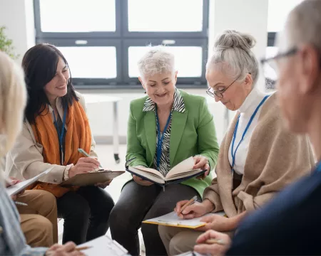 A group of older adults sit together in a bright room, reading and writing in notebooks during a discussion. Two people in the centre look down at an open book.