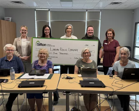 3 members of the Sifton Family Foundation Board along with a facilitator and a London Public Library staff member stand with a large cheque. In front of them, 4 senior students sit with laptops in front of them.