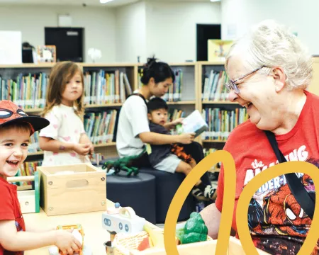 In the foreground an older woman and young child smale and play at a table. In the brackground a woman sits and reads to a child in her lap and a young girl walks by with bookshelves in behind.
