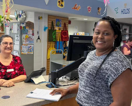 Library staff member stands behind a counter and smiles next to featured patron Shelly at Masonville Branch Library