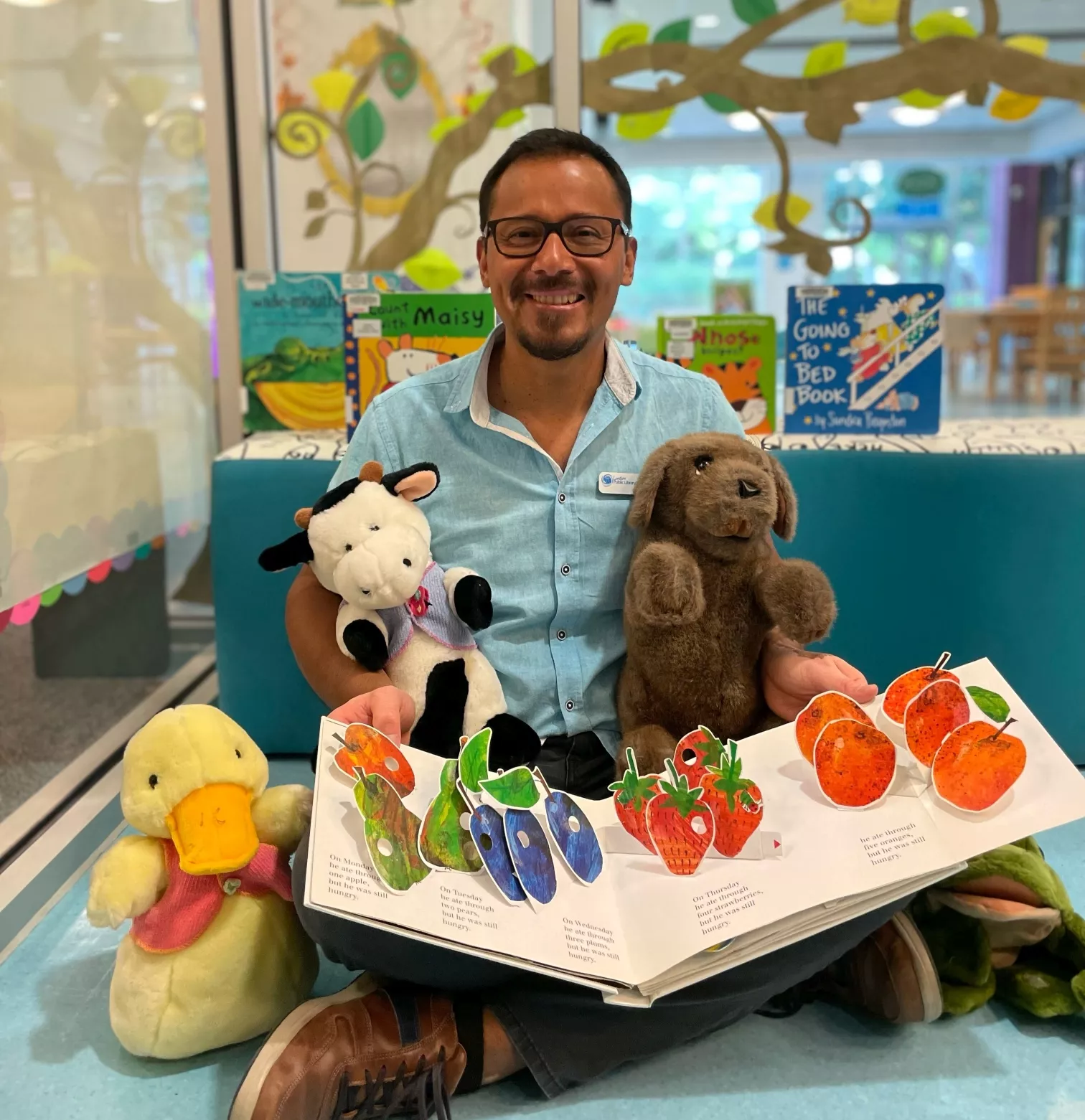 Library staff person sitting on floor in childrens area holding a pop up book in his lap and stuffed animals in his arms