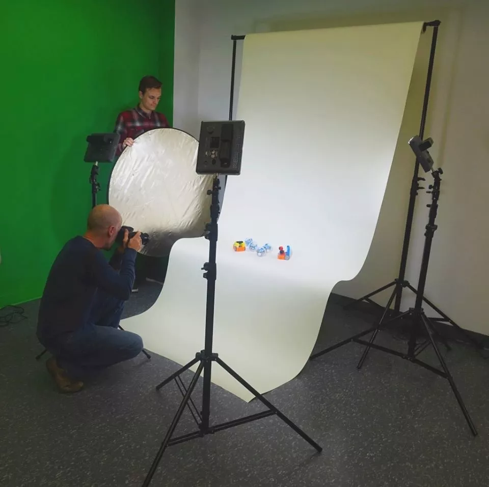 Photo of two men in the Media Lab at Central Library. They are using lights and a white backdrop to take photos of small objects. In the background is a green wall.