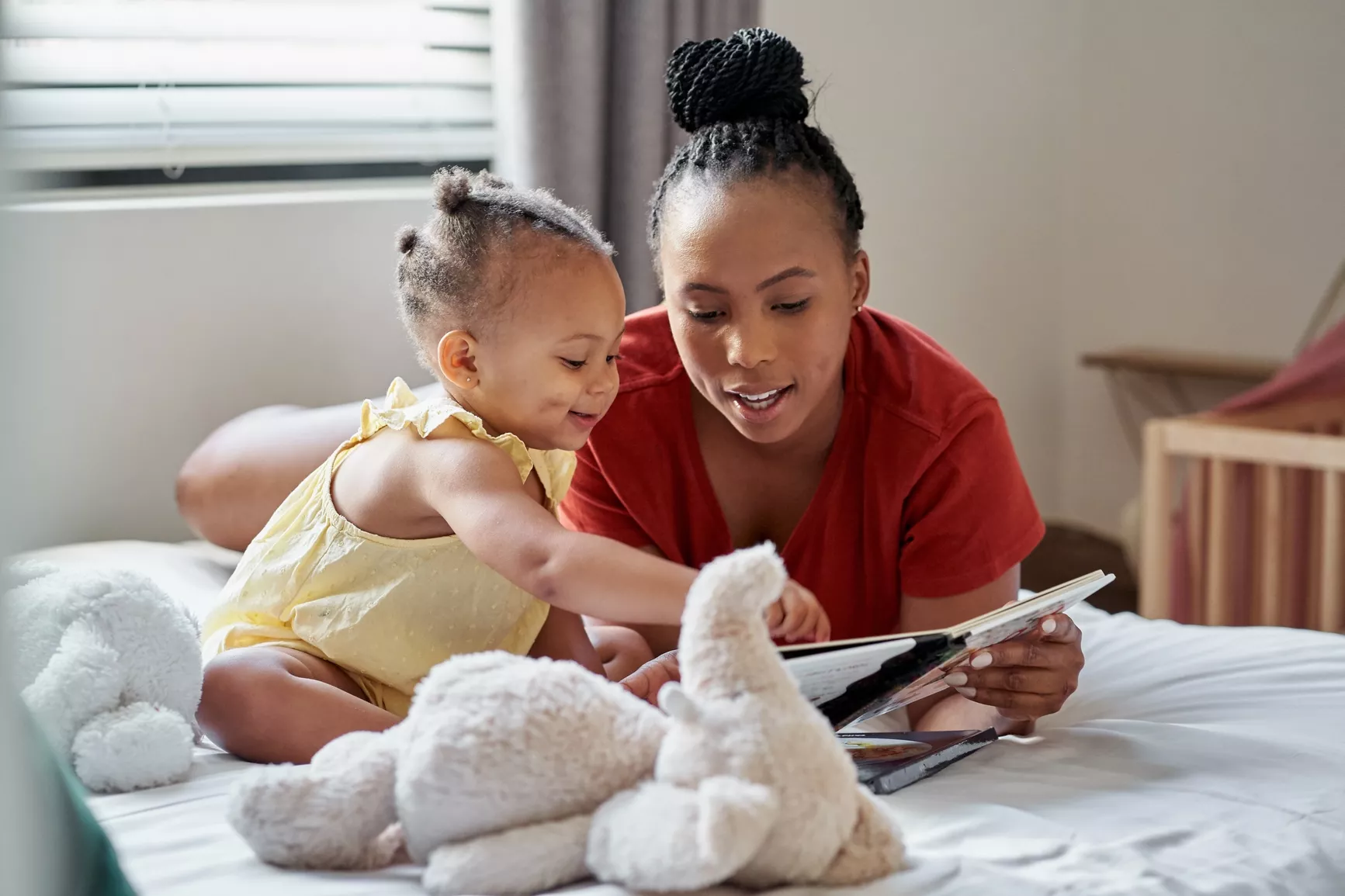 Photo of a woman lying on a bed reading to a young child who is sitting and pointing at the book. Stuffed animals lay on the bed