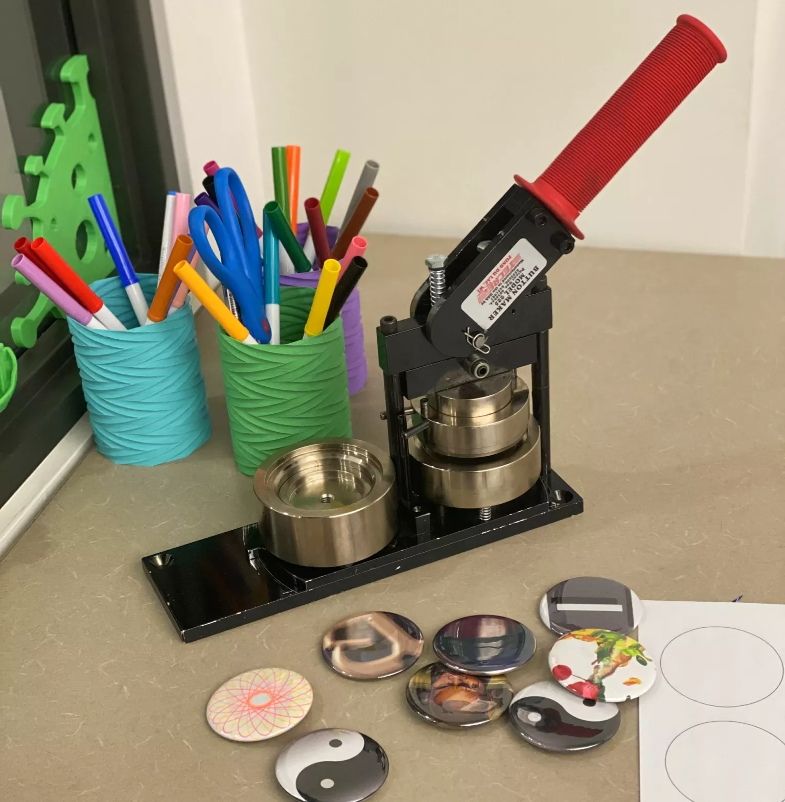 Photo of a button maker on a table with coloured markers in the background and buttons and button templates in the foreground.