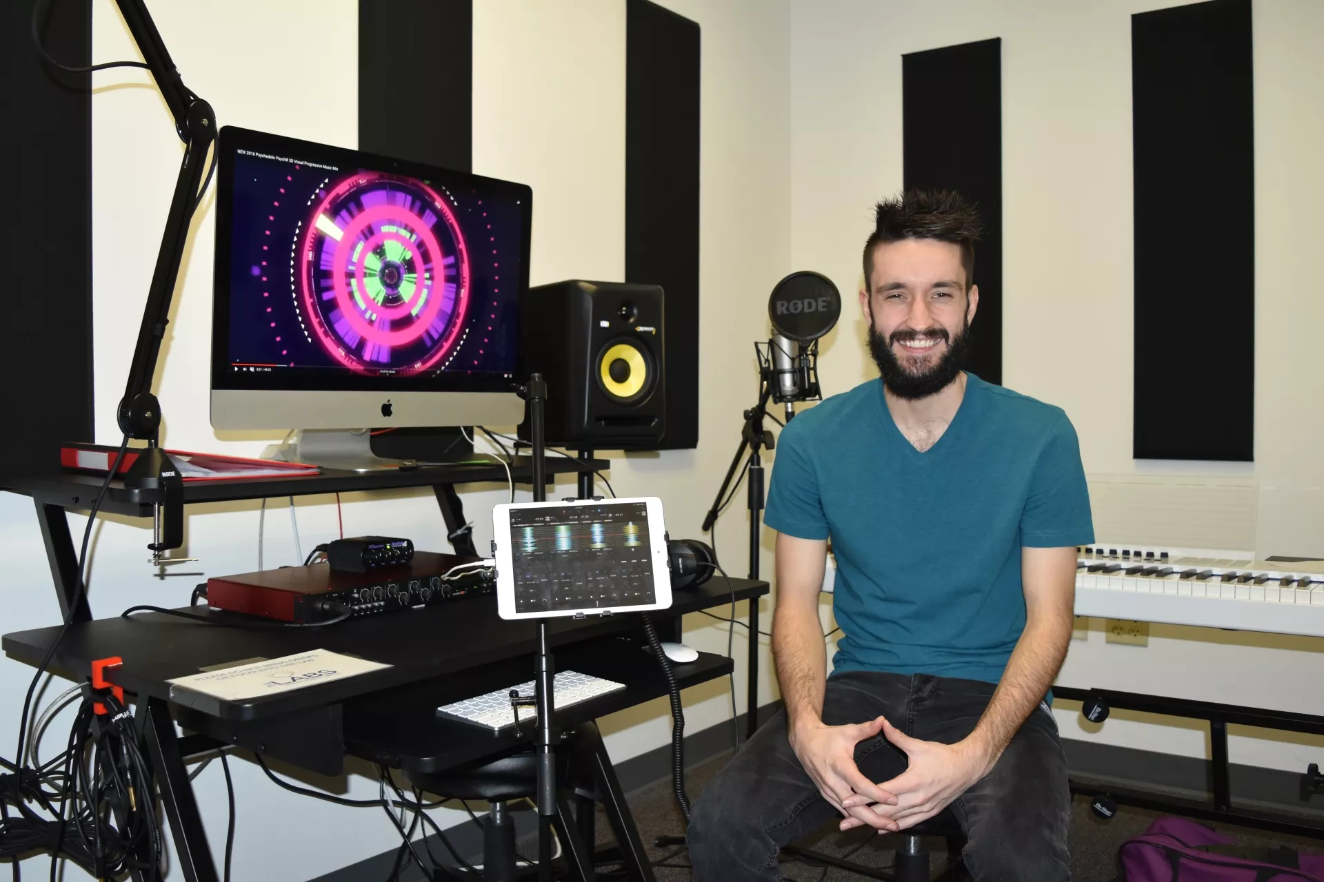 Photo of a smiling man sitting in the audio recording studio. Beside him is a keyboard and monitor. In the back are speakers and in front is a microphone.