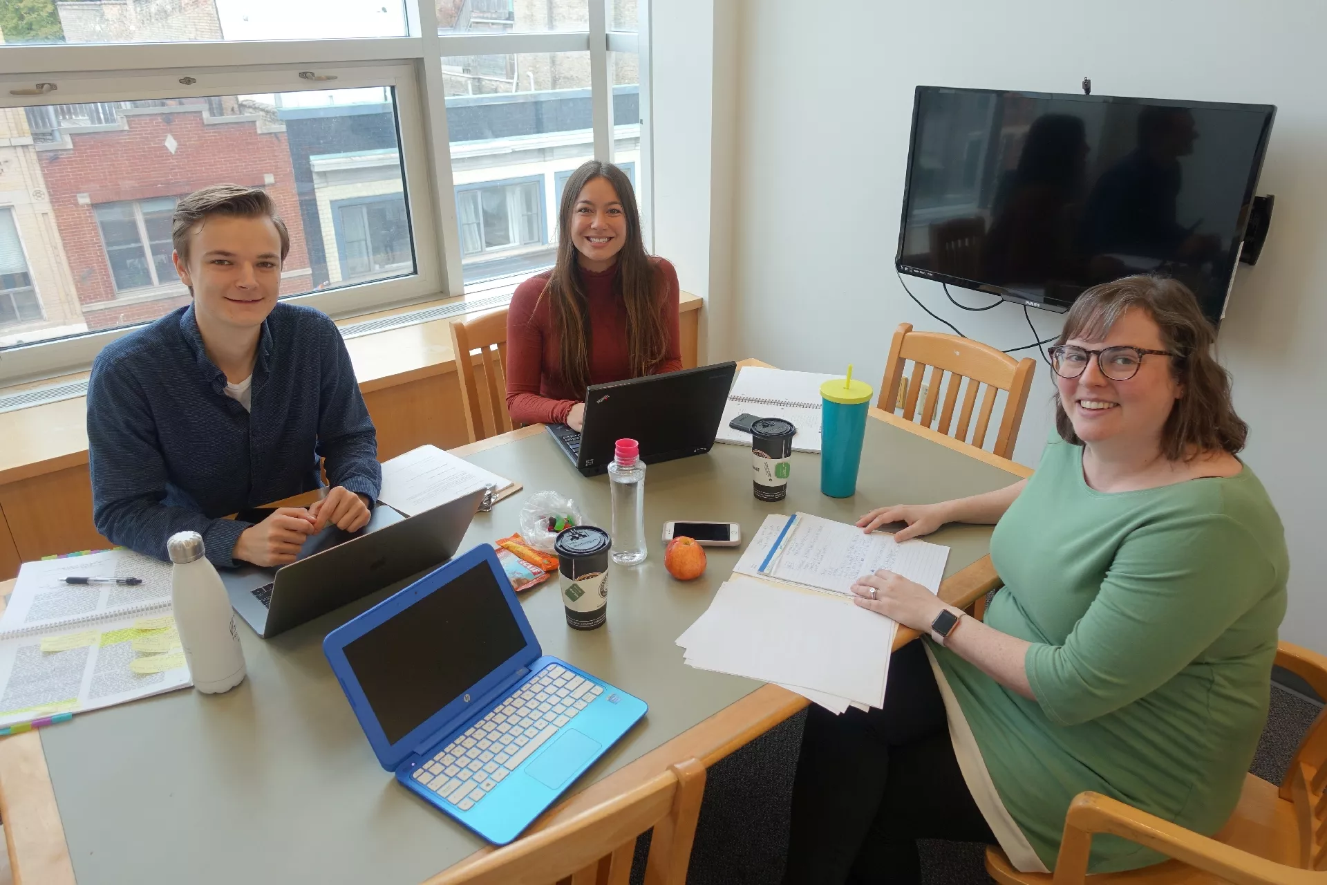 Photo of 3 adults sitting at a table in a study room at Central Library. On the table are laptops notebooks and water bottles. On the wall is a monitor. Large windows look out onto Dundas St.