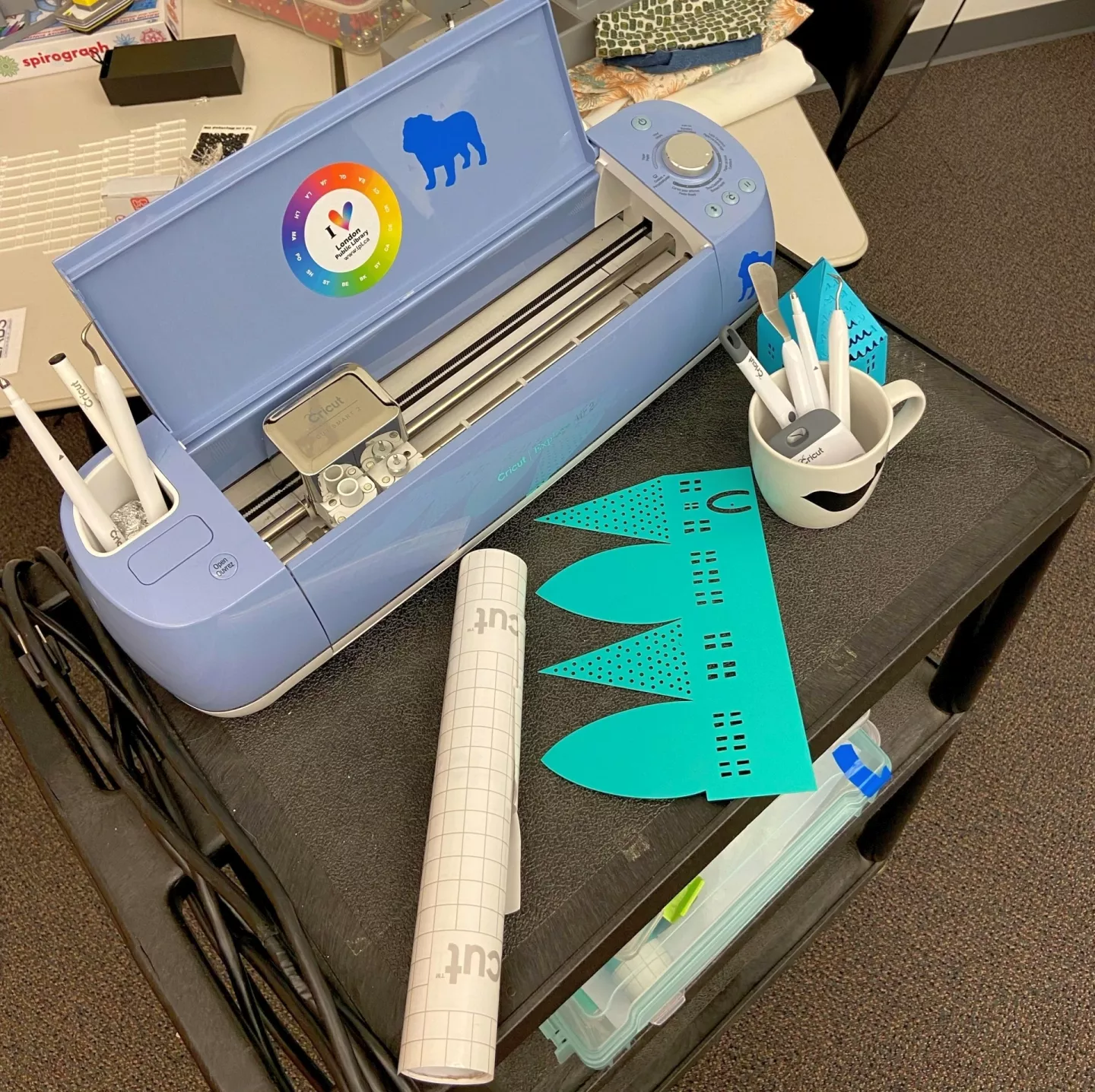 Photo of a blue Cricut 2 machine sitting on a cart. On the card is a mug holding tools, a roll of vinyl and several completed projects.