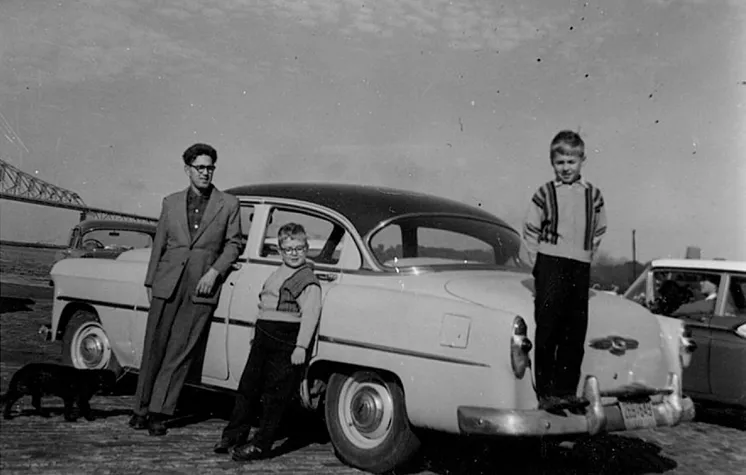 A vintage photo of young Rudy and his family standing beside their car.