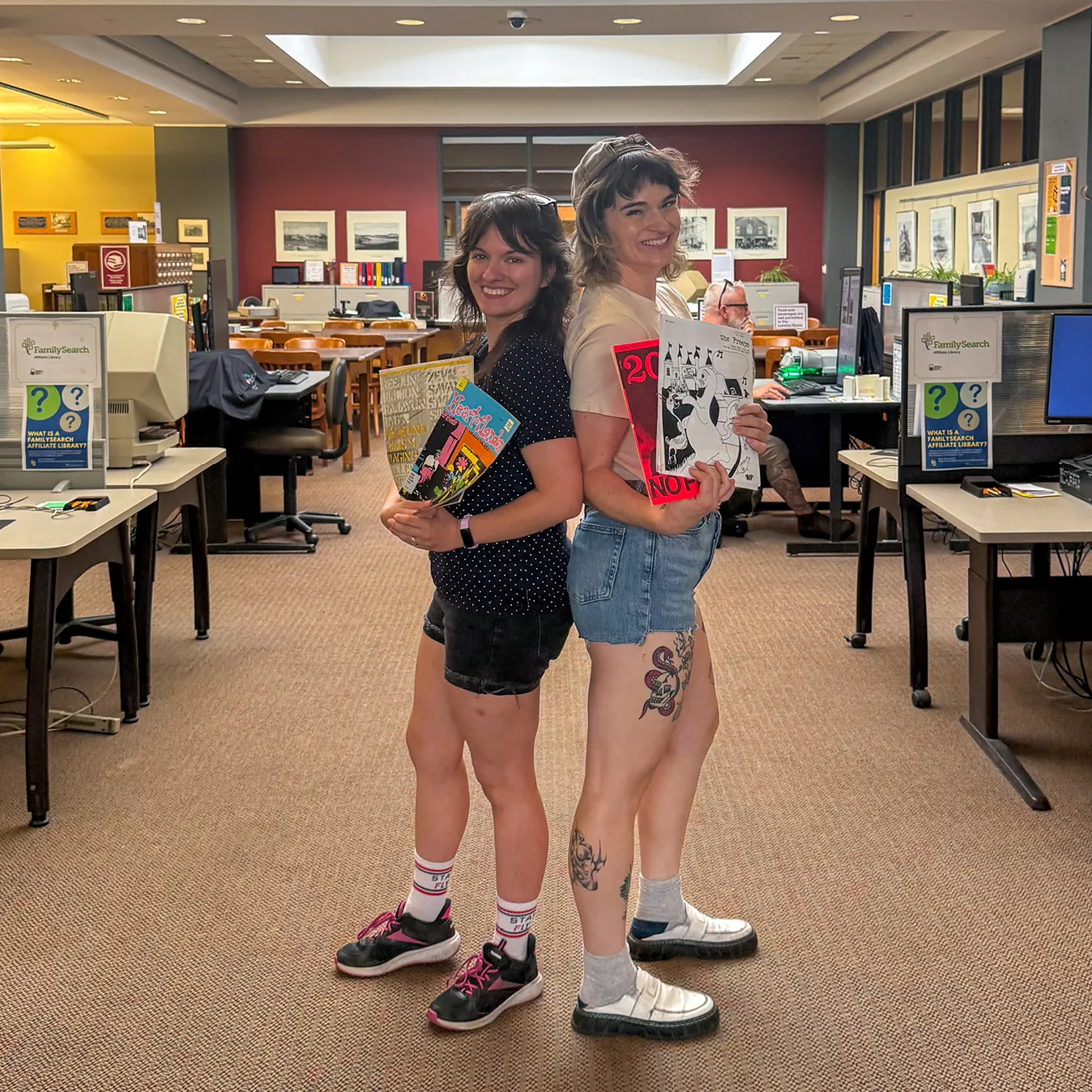 Sammy Orlowski and Jill Clair from Zine Fiends stand back to back in the London Room, holding up zines.