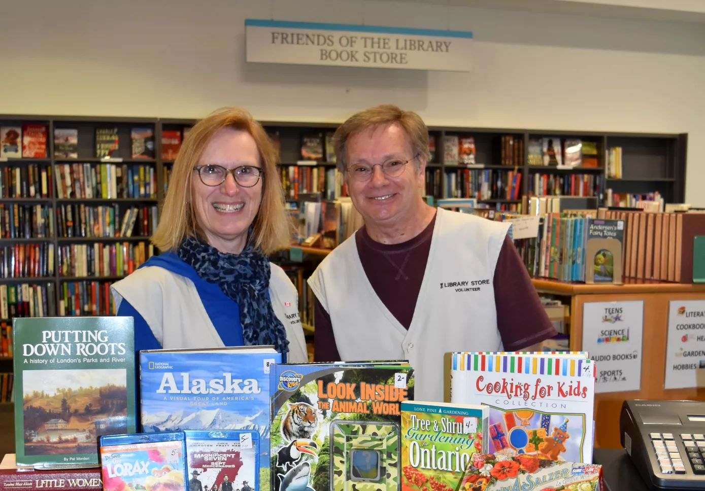 Mary and Scott stand behind a shelf in the Friends Book Store with shelves in the background and books in the foreground.