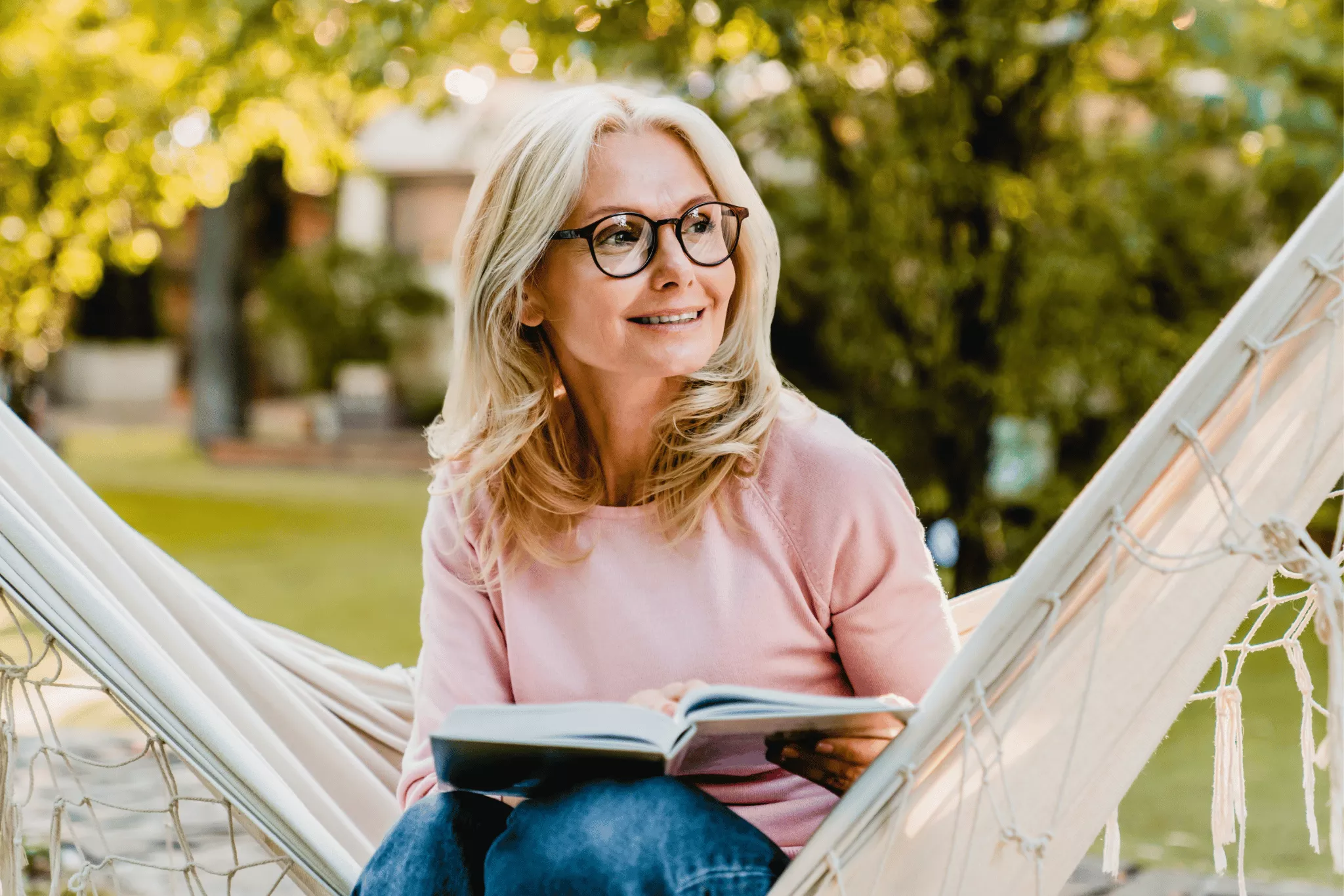 Smiling senior blond woman wearing glasses while reading in hammock in the summer garden