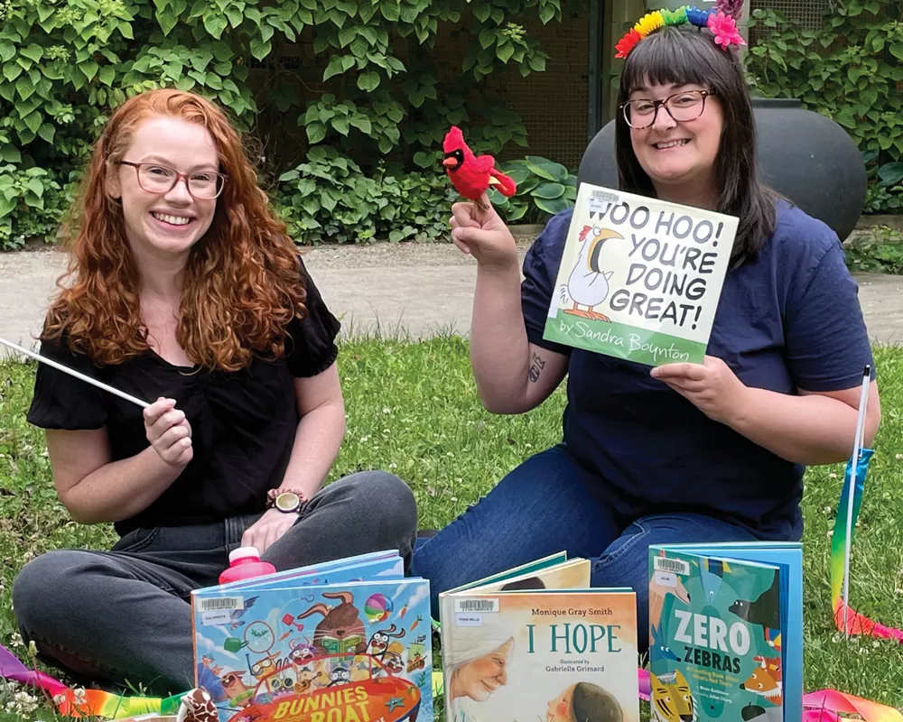 two librarians at an outdoor pop up holding up children's books 