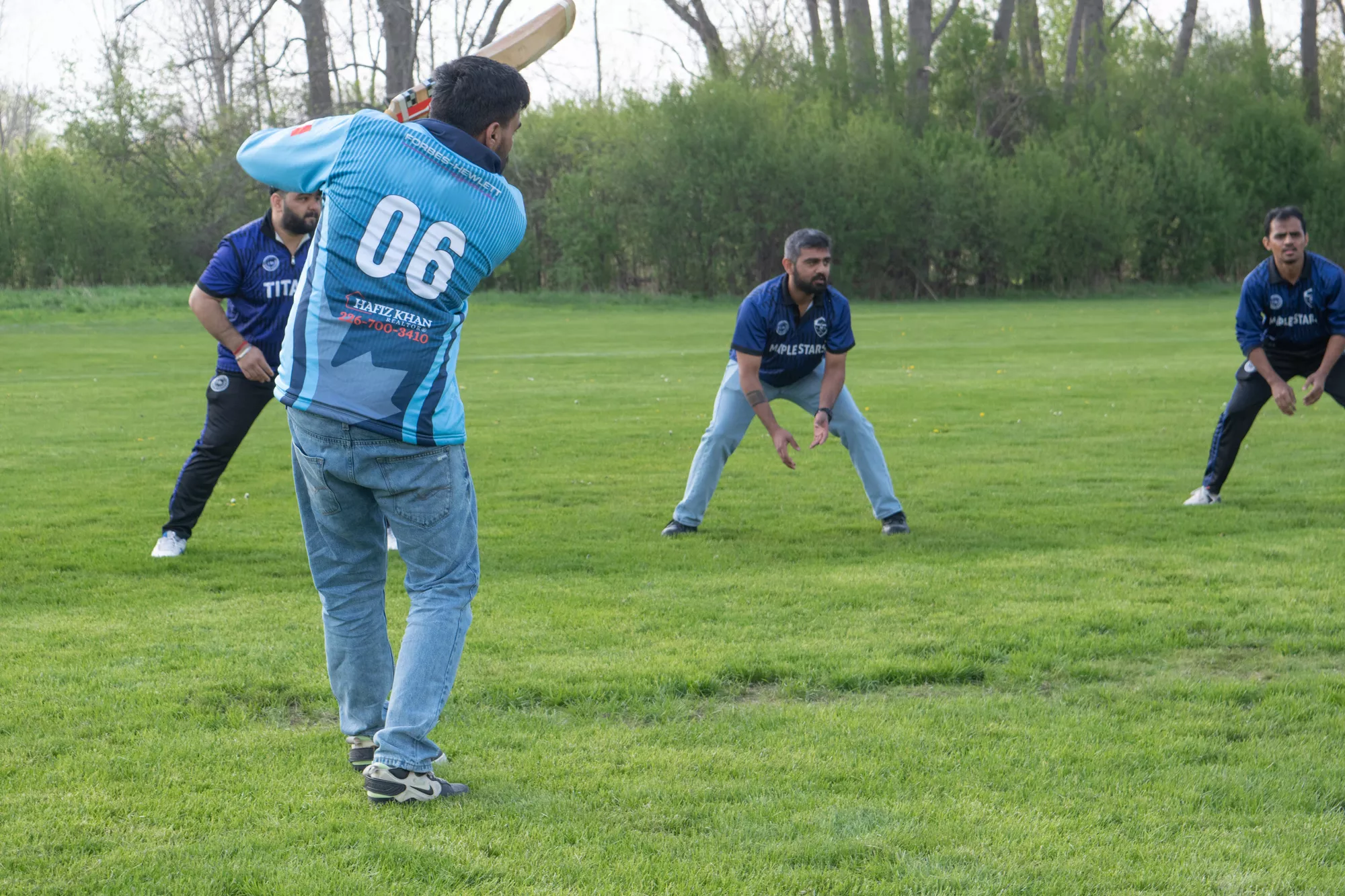 A cricket player swings a bat as three other players crouch nearby. In the background is parkland.