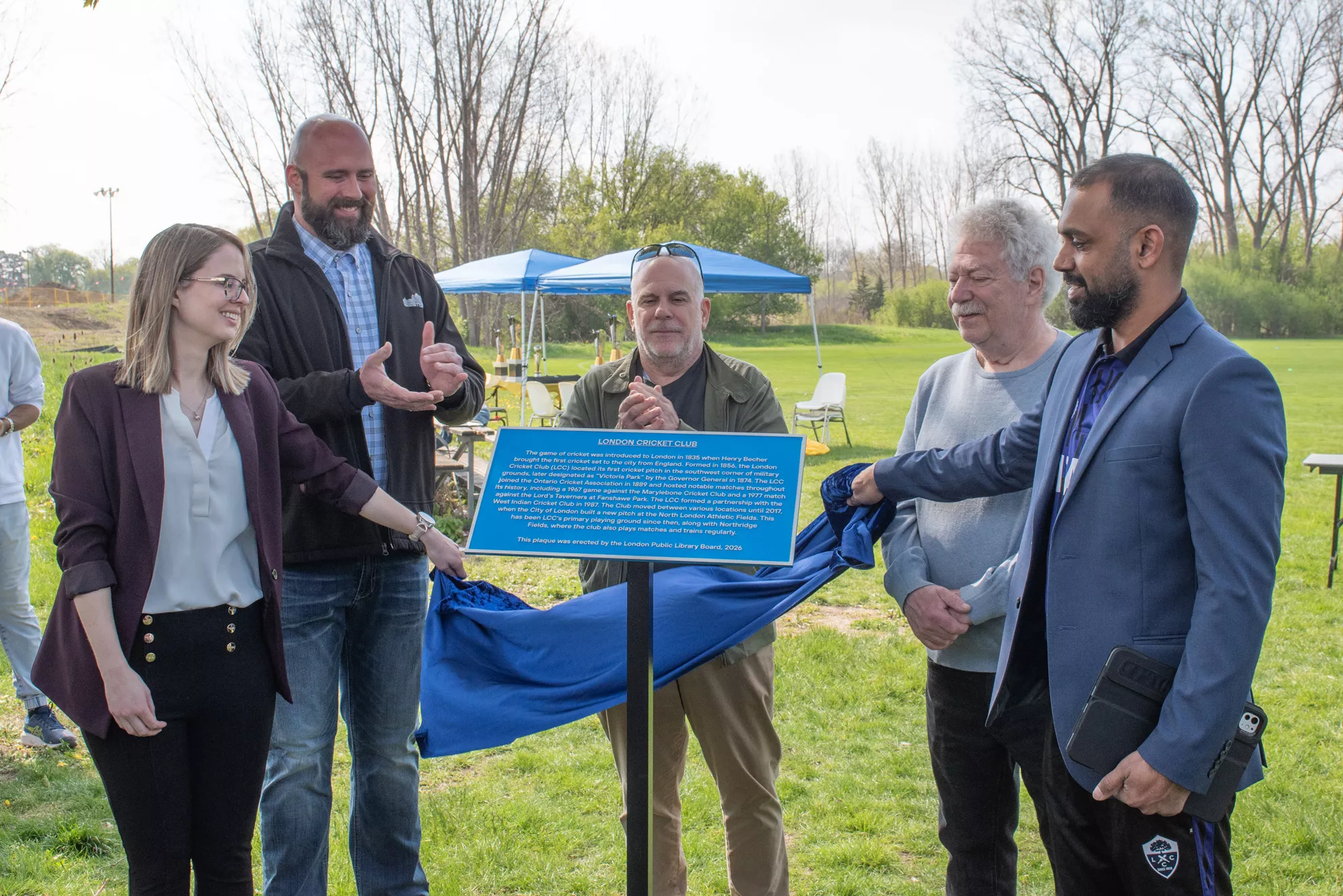 Five people pull a cloth revealing a blue metal plaque on a pole. In the background is parkland and tents.